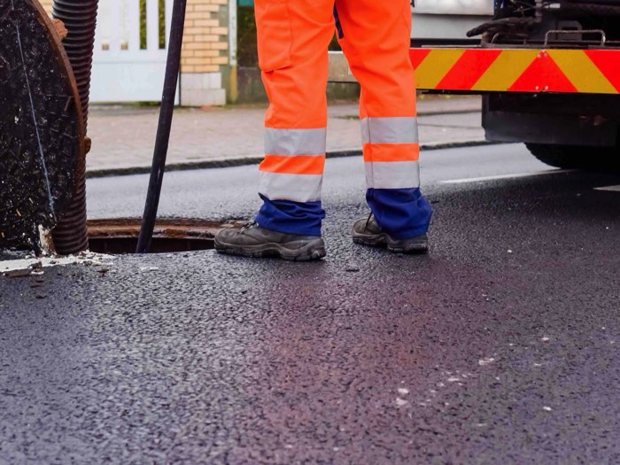 Pracownik ZIĘBUD w pomarańczowym ubraniu ostrzegawczym przy otwartym włazie kanalizacyjnym w jezdni, wąż wprowadzony do studzienki, obok wóz ciśnieniowo-ssący