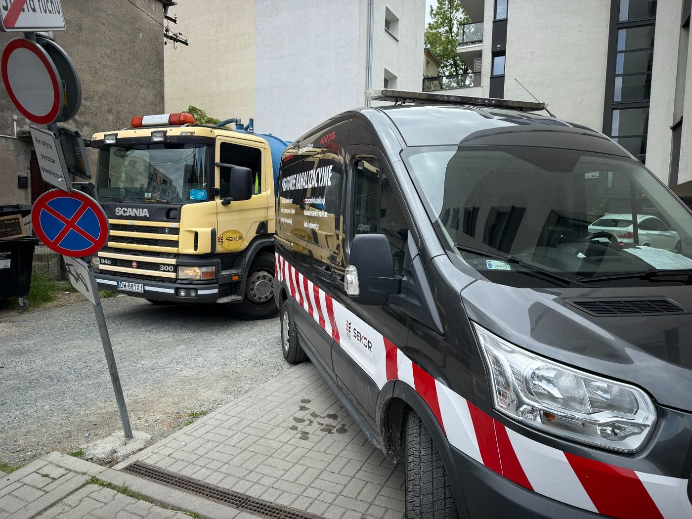 Oznakowany bus serwisowy oraz ciężarówka zaparkowane na osiedlowej drodze przy znakach zakazu